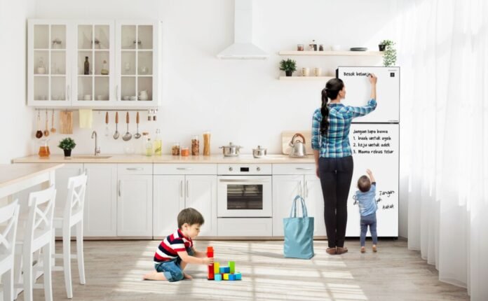 kulkas-dua-pintu-j-tech-inverter-sharp-fitur-tulis-dapur-keluarga Keluarga di dapur modern, anak-anak bermain, sementara ibu menulis daftar belanja di pintu kulkas dua pintu J-TECH Inverter Sharp putih.