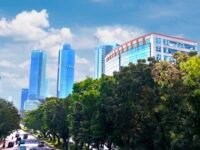 Gedung perkantoran modern dengan fasad kaca biru di sepanjang jalan raya yang ramai dengan kendaraan dan pepohonan hijau yang rindang, properti komersial jakarta