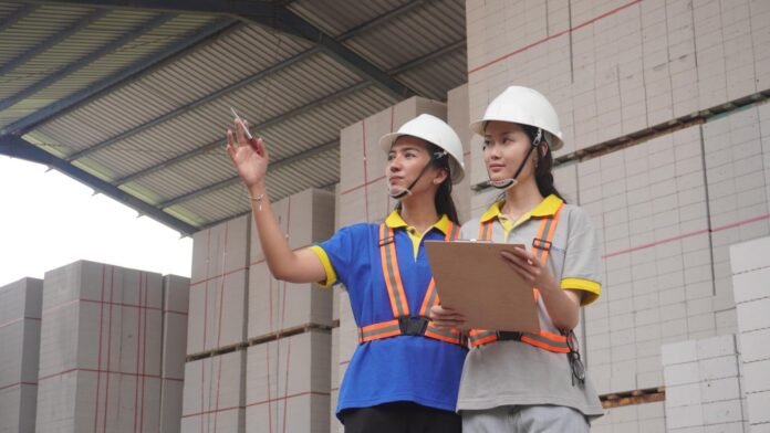 Dua pekerja wanita mengenakan helm keselamatan sedang memeriksa stok bata ringan BLES di area gudang pabrik.