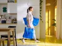 Seorang wanita membawa dua tas IKEA FRAKTA daur ulang berwarna biru berisi barang kebutuhan rumah tangga di dalam dapur