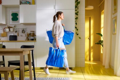 Seorang wanita membawa dua tas IKEA FRAKTA daur ulang berwarna biru berisi barang kebutuhan rumah tangga di dalam dapur