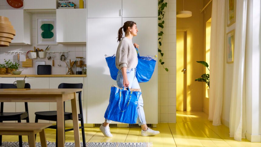 Seorang wanita membawa dua tas IKEA FRAKTA daur ulang berwarna biru berisi barang kebutuhan rumah tangga di dalam dapur